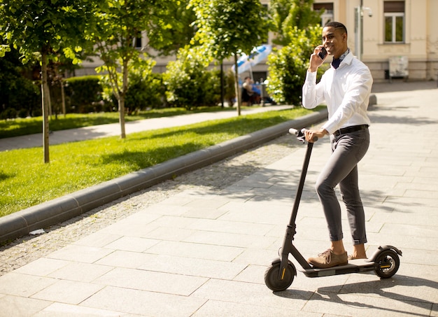 Bel giovane afroamericano che usa il telefono cellulare mentre sta in piedi con uno scooter elettrico su una strada