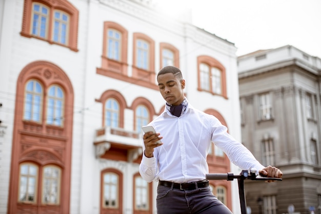 Bel giovane afroamericano che usa il telefono cellulare mentre sta in piedi con uno scooter elettrico su una strada