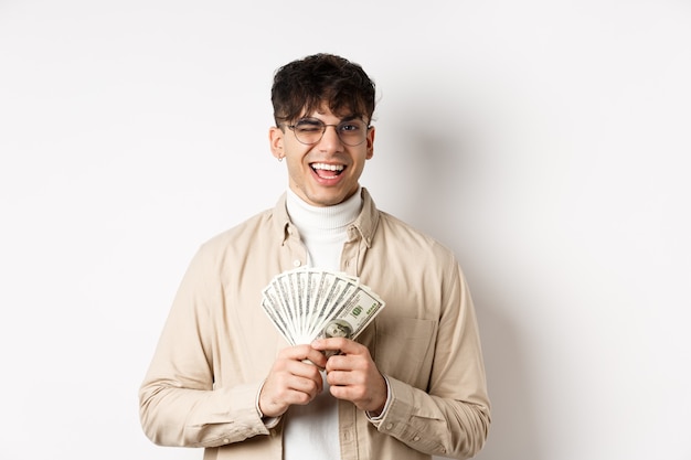 Bel giovane ammiccante e mostrando banconote da un dollaro, tenendo un fan di banconote di denaro e sorridendo compiaciuto, facendo soldi, in piedi sul muro bianco.