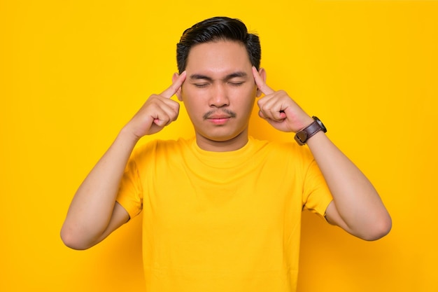 Bel giovane asiatico in maglietta casual che tiene le dita sulle teste cercando di concentrarsi isolato su sfondo giallo Concetto di stile di vita delle persone