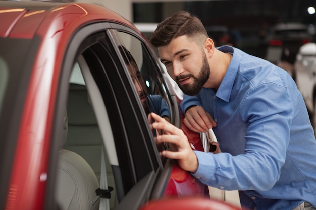 Bel giovane barbuto esaminando nuove auto moderne presso la concessionaria, copia dello spazio