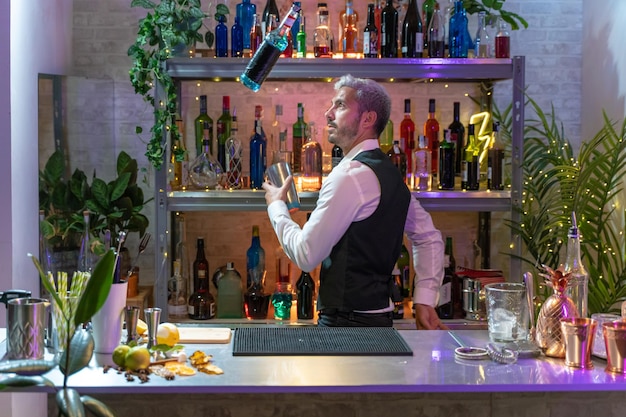 Bel giovane barista all'interno del bar lancia abilmente uno shaker con ghiaccio e una bottiglia in aria Scaffali con bottiglie su sfondo sfocato