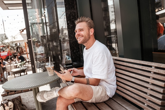 Bel giovane biondo seduto sulla terrazza del caffè con bevanda al caffè sorridente con smartphone mobile su sfondo di riflessione di grande città strada vetro edificio. Hipster millenario in una giornata di sole estivo