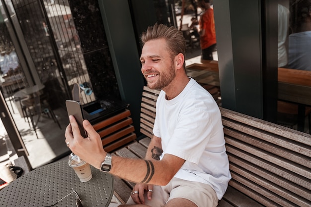 Bel giovane biondo uomo sorridente si siede nel tavolo della terrazza del caffè con la bevanda del caffè che tiene lo smartphone mobile fa selfie, streaming, distacco, simpatia sullo sfondo della costruzione di una strada di grande città.