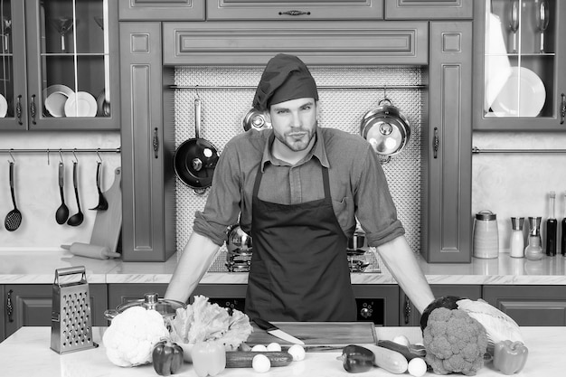 Bel giovane caucasico in grembiule in piedi a tavola con verdure che cucinano a casa preparando il pasto in cucina con superficie in legno piena di stoviglie fantasiose