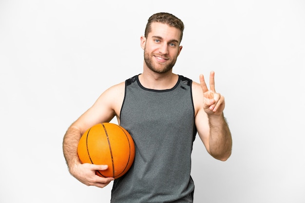 Bel giovane che gioca a basket su sfondo bianco isolato sorridente e mostrando segno di vittoria