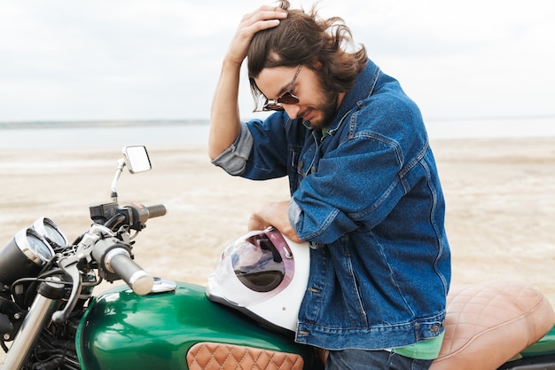 Bel giovane che indossa abiti casual seduto su una moto in spiaggia