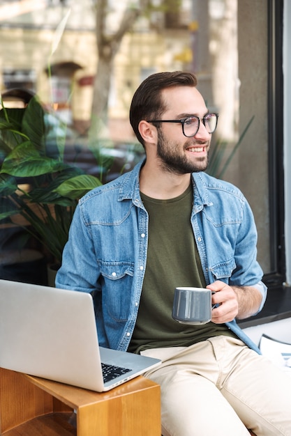 bel giovane che indossa occhiali tenendo una tazza di caffè e digitando sul computer portatile mentre si lavora in un caffè della città all'aperto