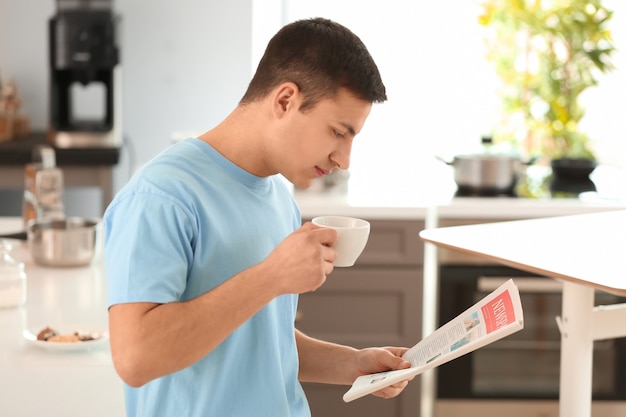 Bel giovane che legge il giornale mentre beve il caffè in cucina