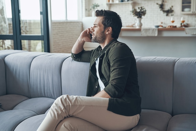 Bel giovane che parla allo smartphone e guarda lontano mentre è seduto sul divano sitting