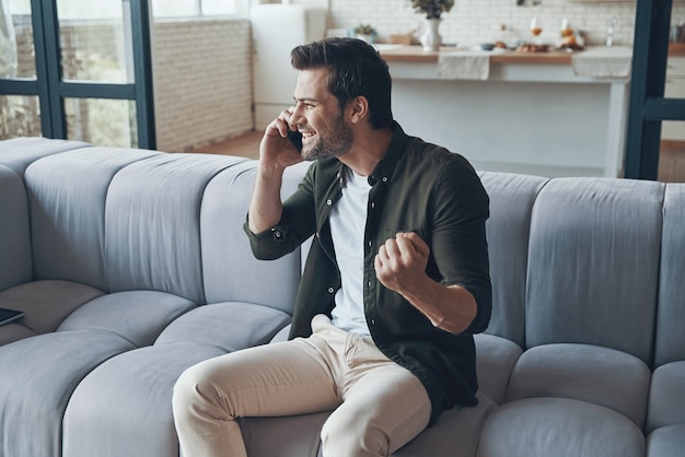 Bel giovane che parla allo smartphone e sorride mentre è seduto sul divano