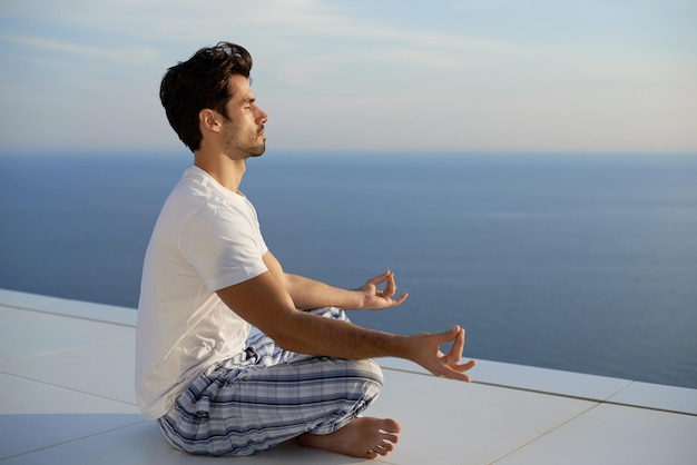 bel giovane che pratica yoga nella moderna terrazza domestica con l'oceano e il tramonto sullo sfondo