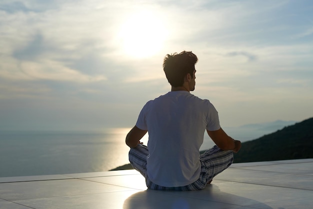 bel giovane che pratica yoga nella moderna terrazza domestica con l'oceano e il tramonto sullo sfondo