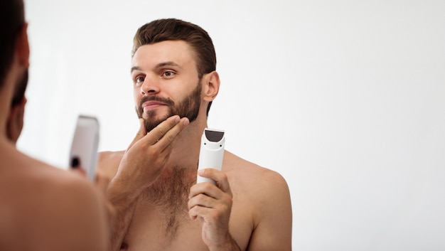 Bel giovane che si rade la barba in bagno. Ritratto di un uomo barbuto nudo alla moda che esamina il suo specchio in casa.