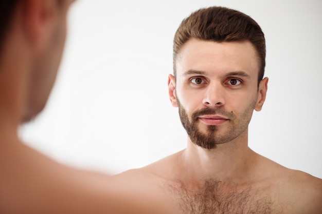 Bel giovane che si rade la barba in bagno. Ritratto di un uomo barbuto nudo alla moda che esamina il suo specchio in casa.