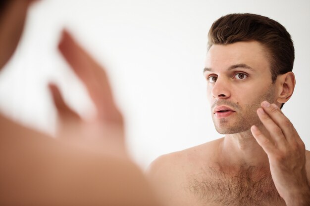 Bel giovane che si rade la barba in bagno. Ritratto di un uomo barbuto nudo alla moda che esamina il suo specchio in casa.