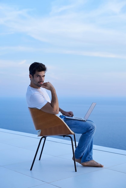bel giovane che si rilassa e lavora al computer portatile sul balcone di casa mentre guarda il tramonto