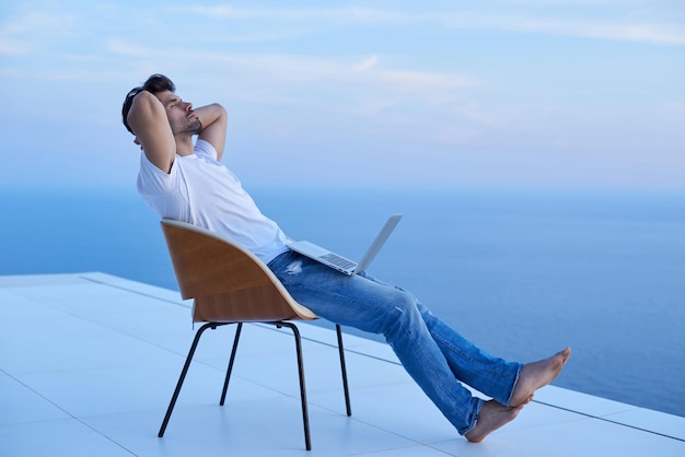 bel giovane che si rilassa e lavora al computer portatile sul balcone di casa mentre guarda il tramonto