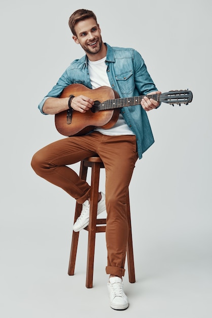 Bel giovane che sorride e suona la chitarra mentre è seduto su uno sfondo grigio