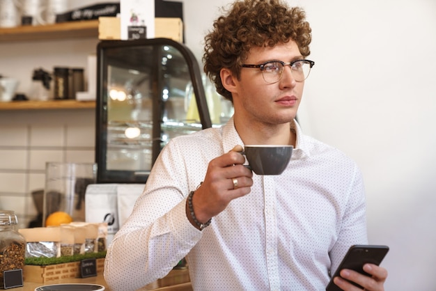 Bel giovane che utilizza il telefono cellulare mentre beve caffè al bar al chiuso