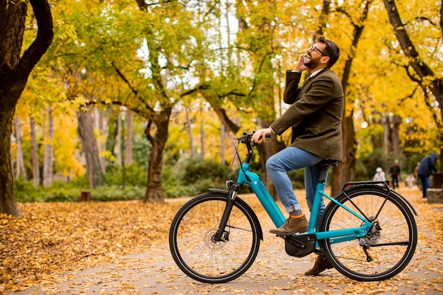 Bel giovane che utilizza il telefono cellulare sulla bicicletta elettrica al parco autunnale