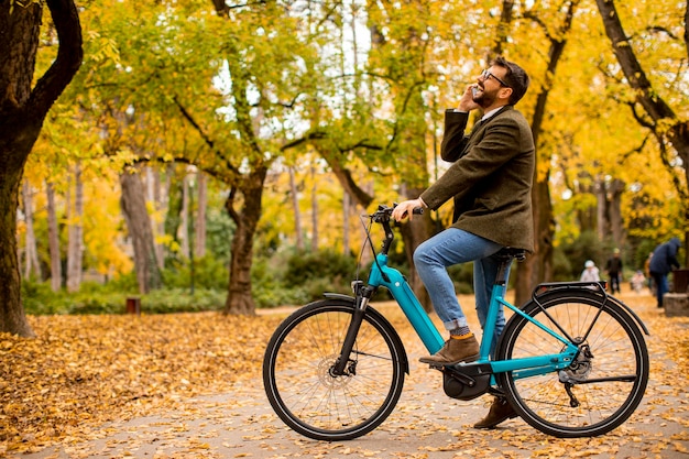 Bel giovane che utilizza il telefono cellulare sulla bicicletta elettrica al parco autunnale