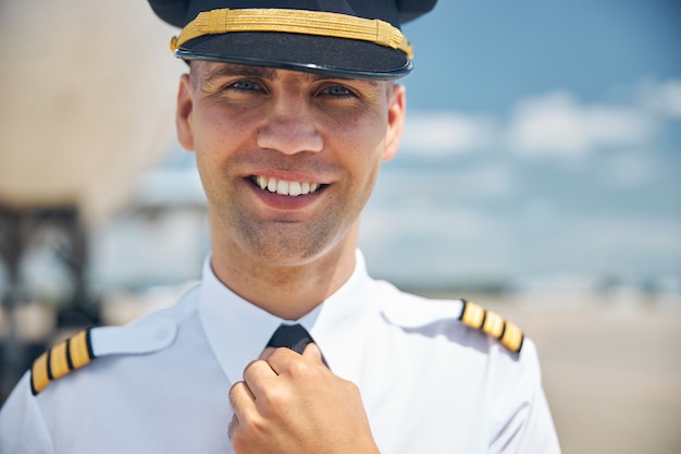 Bel giovane con cappello da capitano che guarda la telecamera e sorride mentre si aggiusta la cravatta