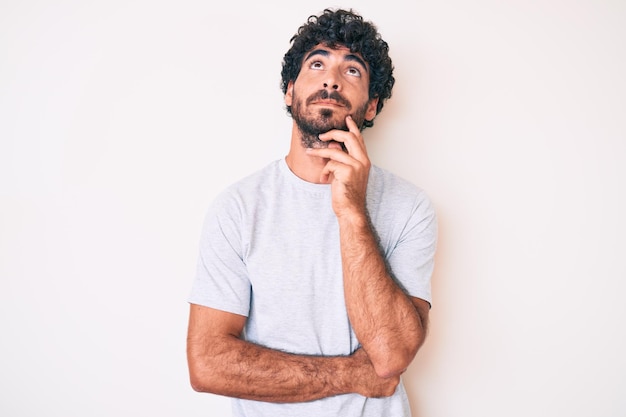 Bel giovane con i capelli ricci e l'orso che indossa una maglietta casual faccia seria pensando alla domanda con la mano sul mento, pensieroso sull'idea confusa