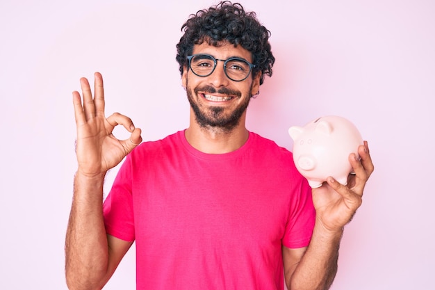 Bel giovane con i capelli ricci e l'orso che tiene salvadanaio facendo segno ok con le dita sorridenti gesti amichevoli simbolo eccellente