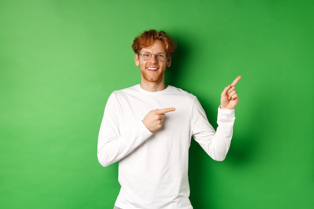 Bel giovane con i capelli rossi e gli occhiali, puntando il dito a destra nello spazio della copia e sorridente, in piedi su sfondo verde.