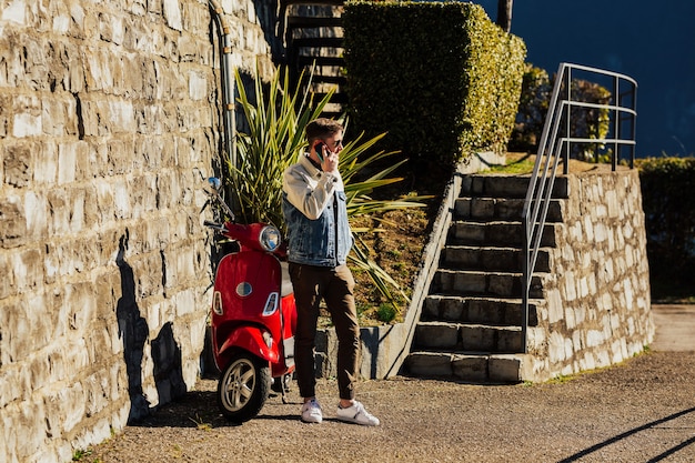 Bel giovane con il telefono cellulare in piedi vicino alla moto rossa in città e parlare.