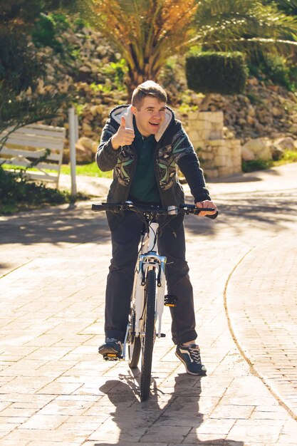 Bel giovane con la bicicletta che mostra i pollici in su nel parco in giornata di sole