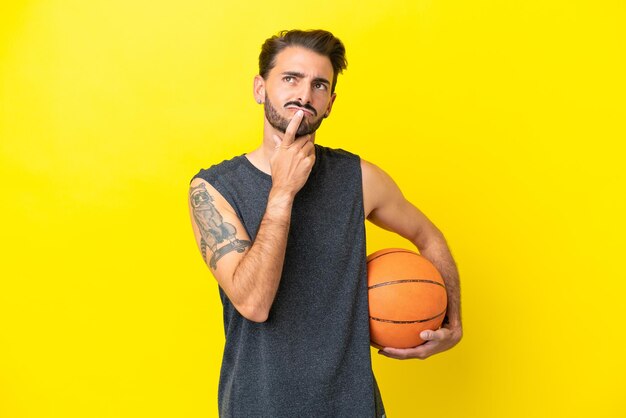 Bel giovane giocatore di basket uomo isolato su sfondo giallo che ha dubbi mentre cerca