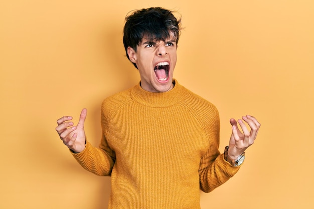 Bel giovane hipster che indossa un maglione giallo casual pazzo e pazzo urlando e urlando con espressione aggressiva e braccia alzate concetto di frustrazione