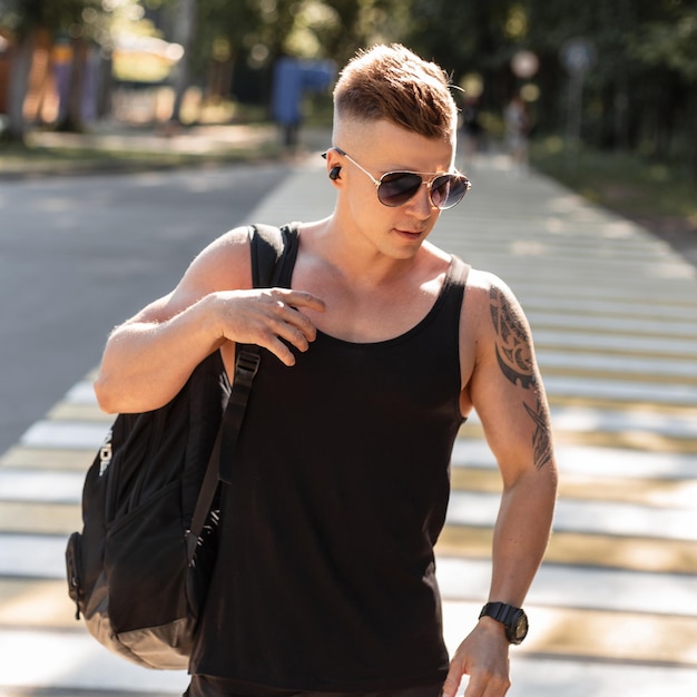 Bel giovane hipster in abiti estivi di moda nera con canotta e pantaloncini con zaino cammina per strada e ascolta musica