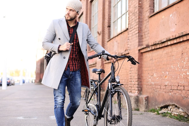 Bel giovane in cappotto grigio e cappello che spinge la sua bici lungo una strada della città.