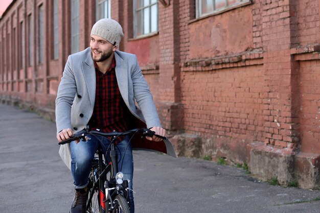 Bel giovane in cappotto grigio e cappello in sella a una strada in bicicletta in città.