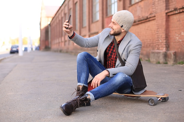 Bel giovane in cappotto grigio e cappello seduto sul longboard e prendendo selfie per strada in città.