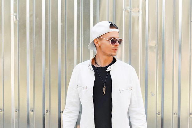 Bel giovane in una giacca alla moda con un berretto da baseball bianco in occhiali da sole vicino al muro di metallo