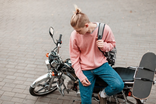 Bel giovane in vestiti alla moda con uno zaino e con uno skateboard siede una moto