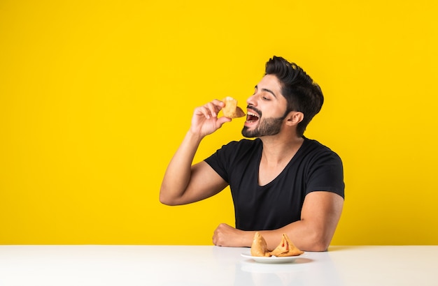 Bel giovane indiano barbuto che mangia spuntini Samosa mentre è seduto al tavolo bianco su sfondo giallo
