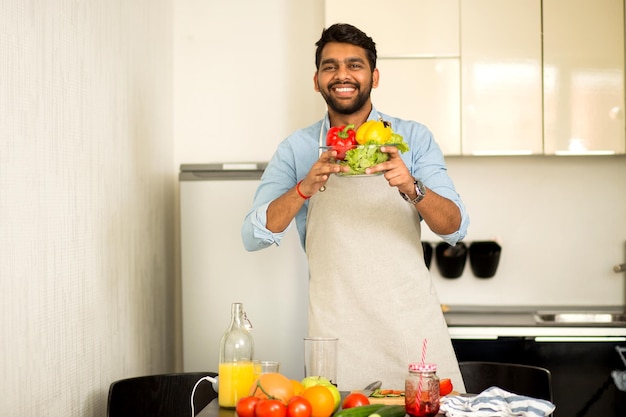 Bel giovane indiano in camicia blu e grembiule che guarda l'obbiettivo e sorride mentre si trova in cucina a casa, preparando insalata di verdure, prendendosi cura della sua salute. Cibo sano, concetto di cucina.