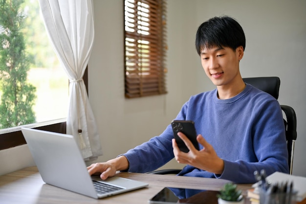 Bel giovane lavoratore d'ufficio maschio asiatico che utilizza il suo smartphone alla scrivania dell'ufficio