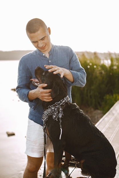 Bel giovane maschio in abito casual che gioca con un cane carino mentre si trova vicino al lago. Ragazzo che indossa una maglietta blu e pantaloncini di jeans bianchi