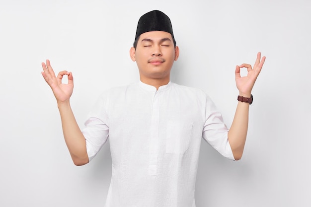 Bel giovane musulmano asiatico in piedi con gli occhi chiusi che si concentra e si rilassa mentre medita isolato su sfondo bianco Concetto di stile di vita islamico religioso delle persone