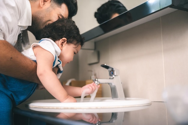 Bel giovane padre che solleva la piccola figlia mentre la istruisce sulla pulizia aiutandola a lavarsi le mani sotto l'acqua corrente del rubinetto nel lavabo del bagno