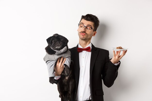 Bel giovane proprietario di cane in abito elegante che tiene in mano un simpatico carlino nero e un piatto con cibo per animali, in piedi su sfondo bianco