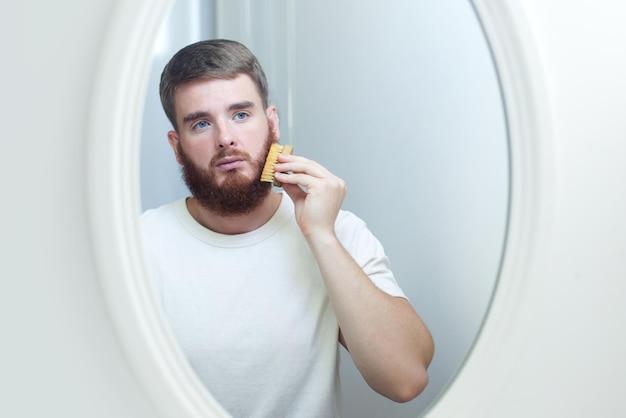 Bel giovane ragazzo barbuto europeo brutale con i capelli della barba sta pettinando la barba sul viso con a