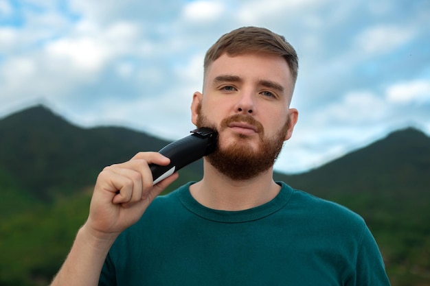 Bel giovane ragazzo barbuto L'uomo brutale europeo con i capelli della barba si sta radendo la barba sul viso con un rifinitore Accessorio per prodotti per la cura personale maschile sulla natura Sfondo estivo naturale