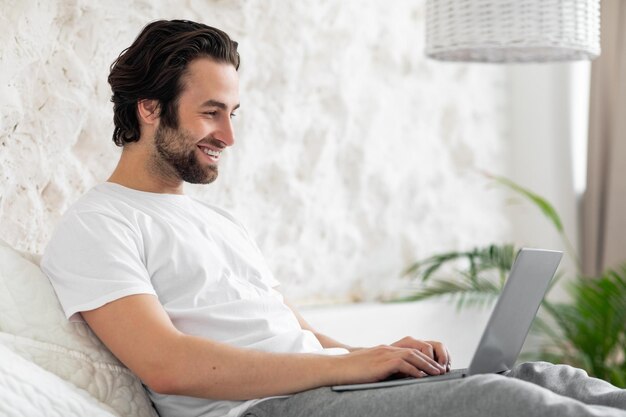 Bel giovane ragazzo che usa il laptop a letto lavorando da casa